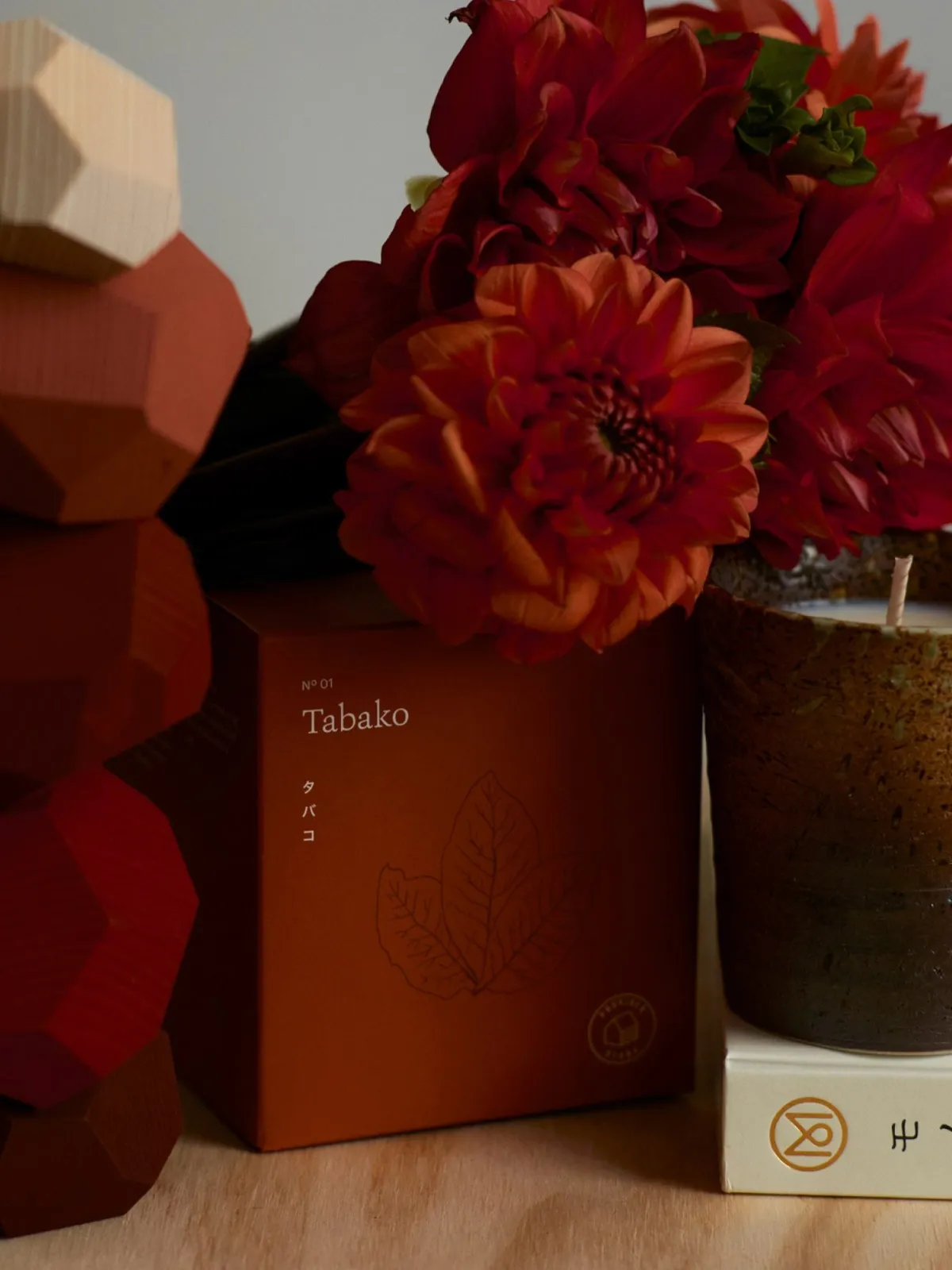 A styled still life with dahlias, a Tabako candle box, and a dark ceramic candle.