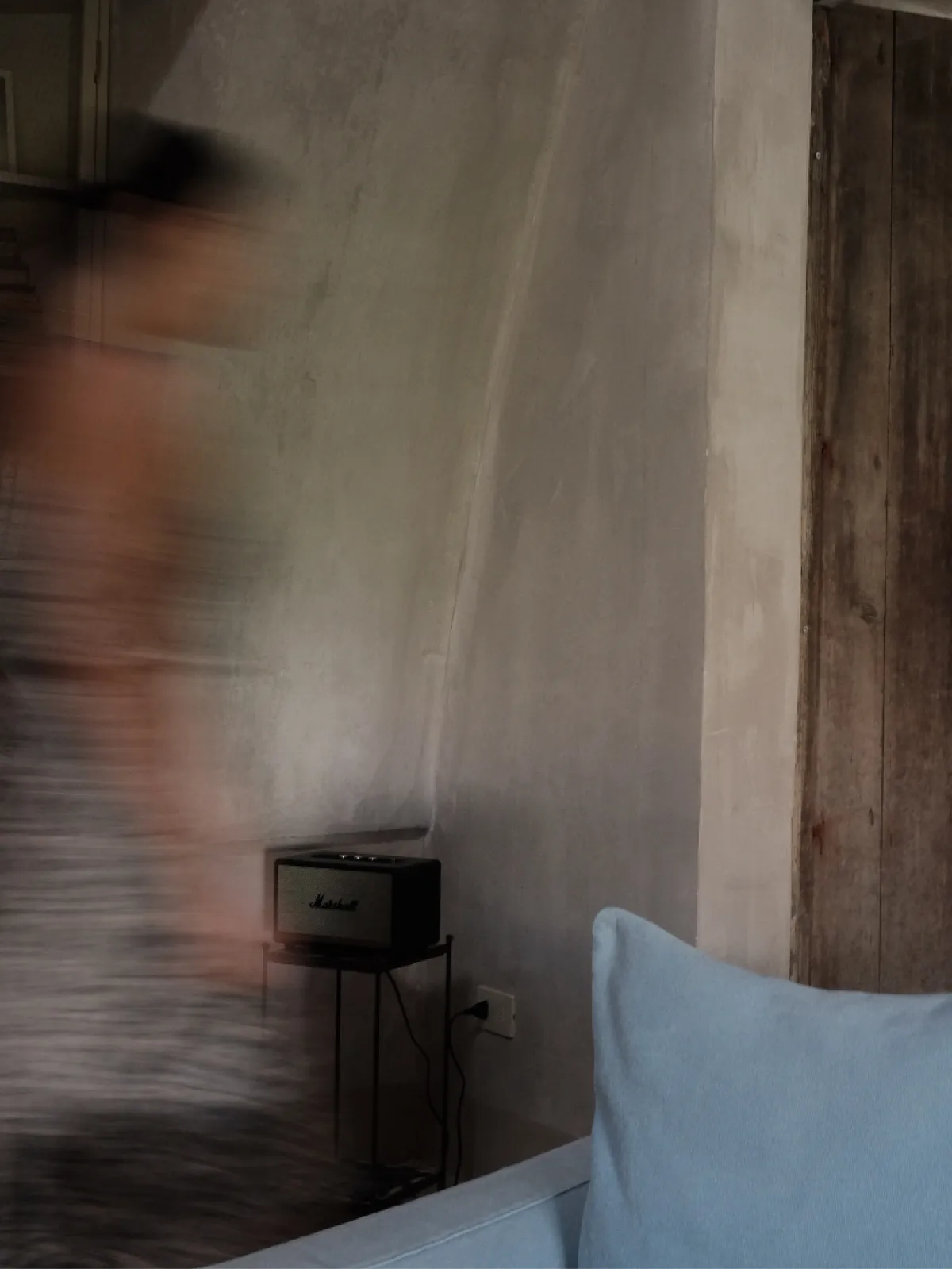 A blurred figure walking past a textured wall beside a pale blue cushion.